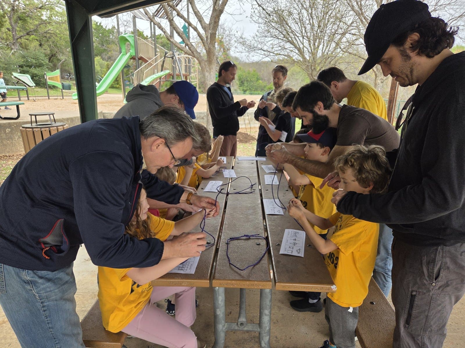 Scouts and adult leaders working together on a hands-on project at picnic tables in an outdoor park pavilion