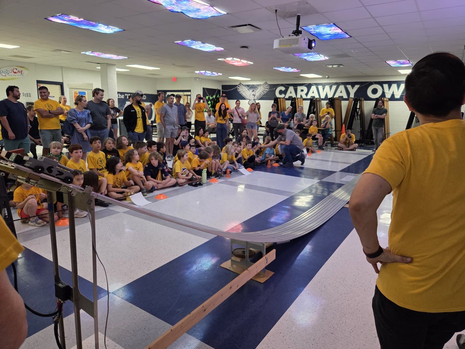 A crowd of scouts and families gathered around a Pinewood Derby race track set up in the school cafeteria