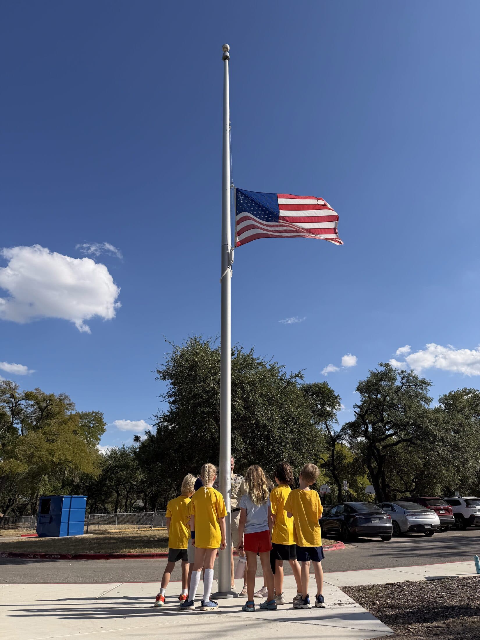 Pack 162 scouts standing at the flagpole at Kathy Caraway Elementary, watching the flag fly against a blue Texas sky