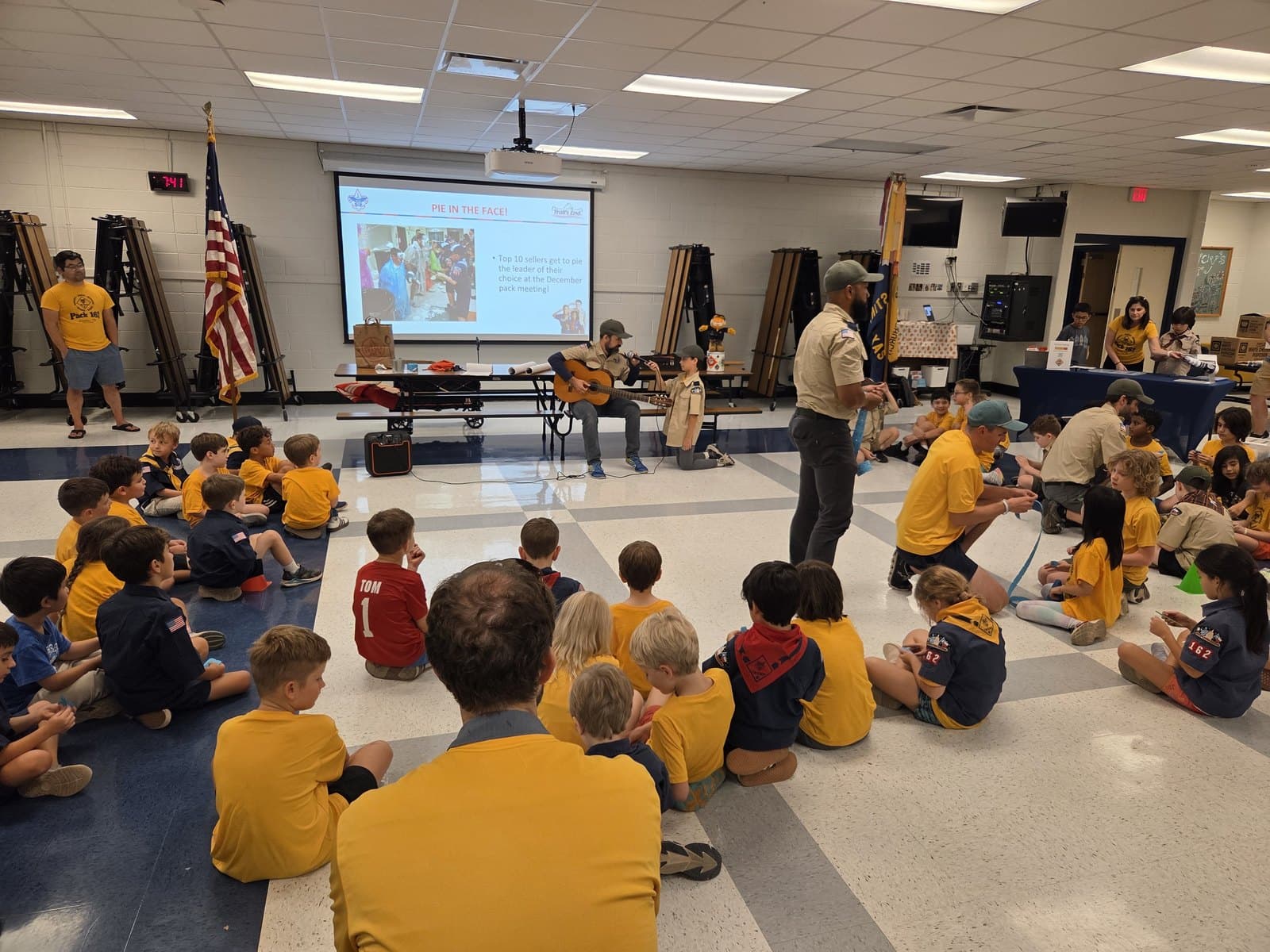 A full Cub Scout pack seated on a cafeteria floor watching a performance during a pack meeting