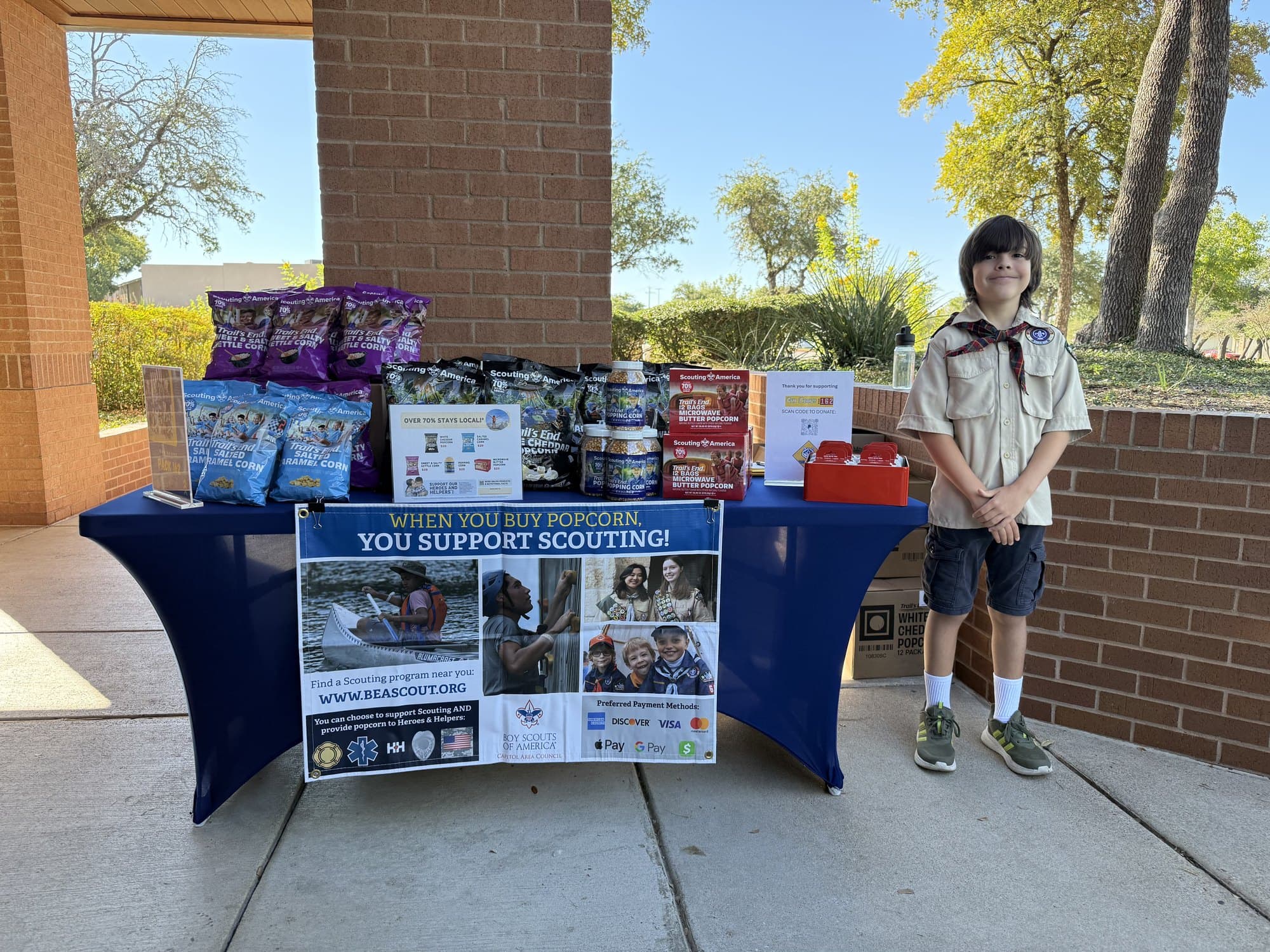 A Cub Scout standing beside a fully stocked popcorn fundraiser table under a covered walkway