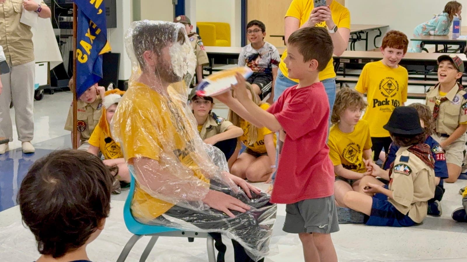 A Cub Scout pushing a pie into a seated leader's face at a pack meeting, watched by the rest of the pack