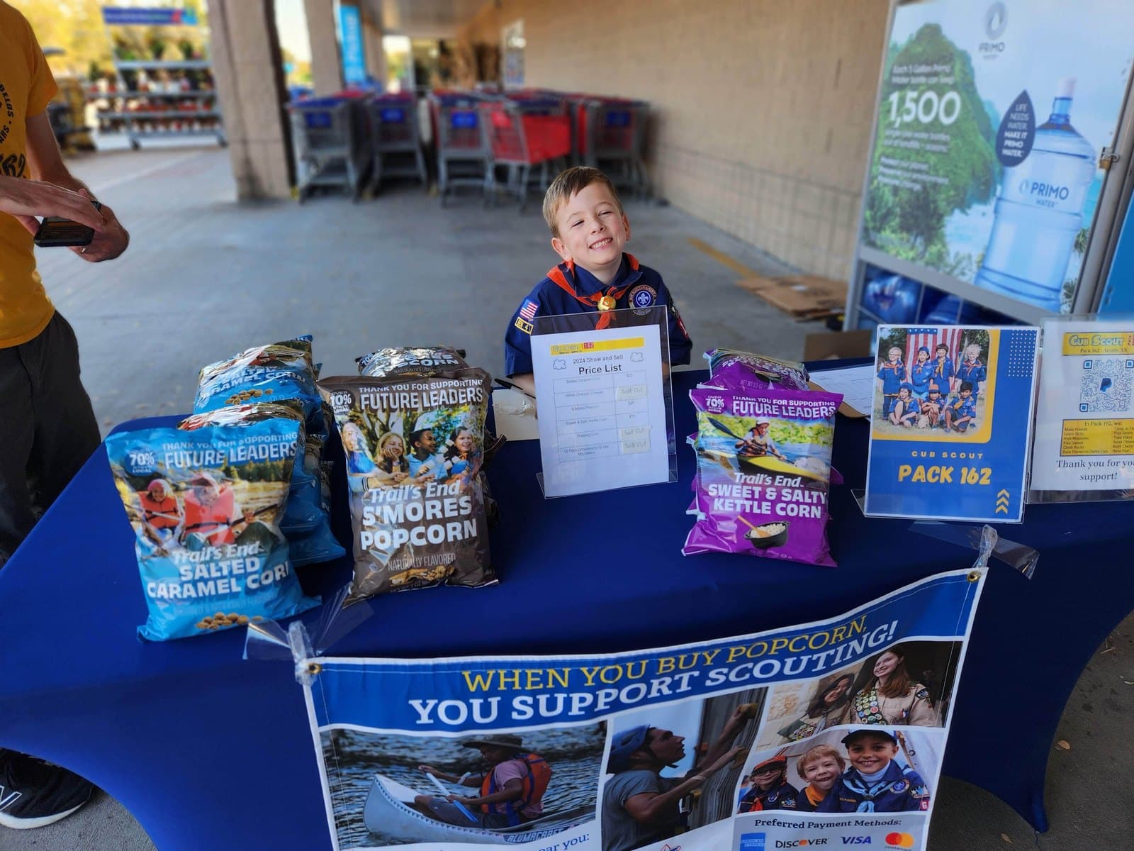 A Cub Scout in uniform smiling behind a table of popcorn for sale outside a store, with a Pack 162 sign