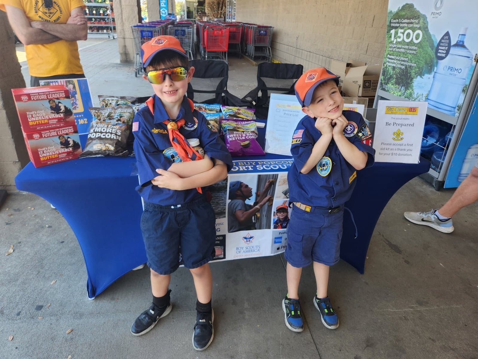 Two young Cub Scouts in uniform with orange caps smiling together at a popcorn table outside a store