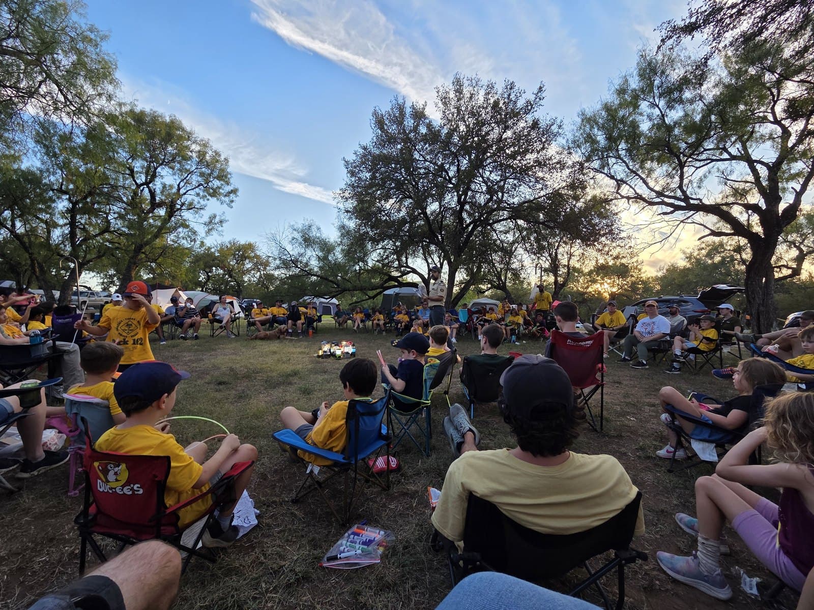 A large circle of scouts and family members sitting in camp chairs under trees at dusk during a fall campout