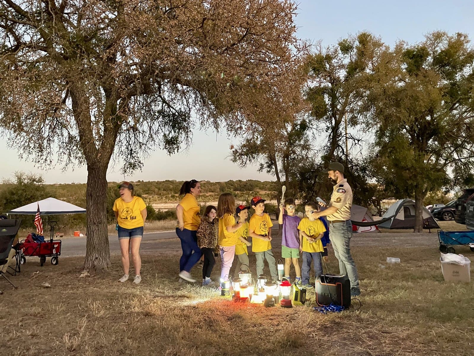 A family gathered around glowing camp lanterns at dusk, tents visible in the background