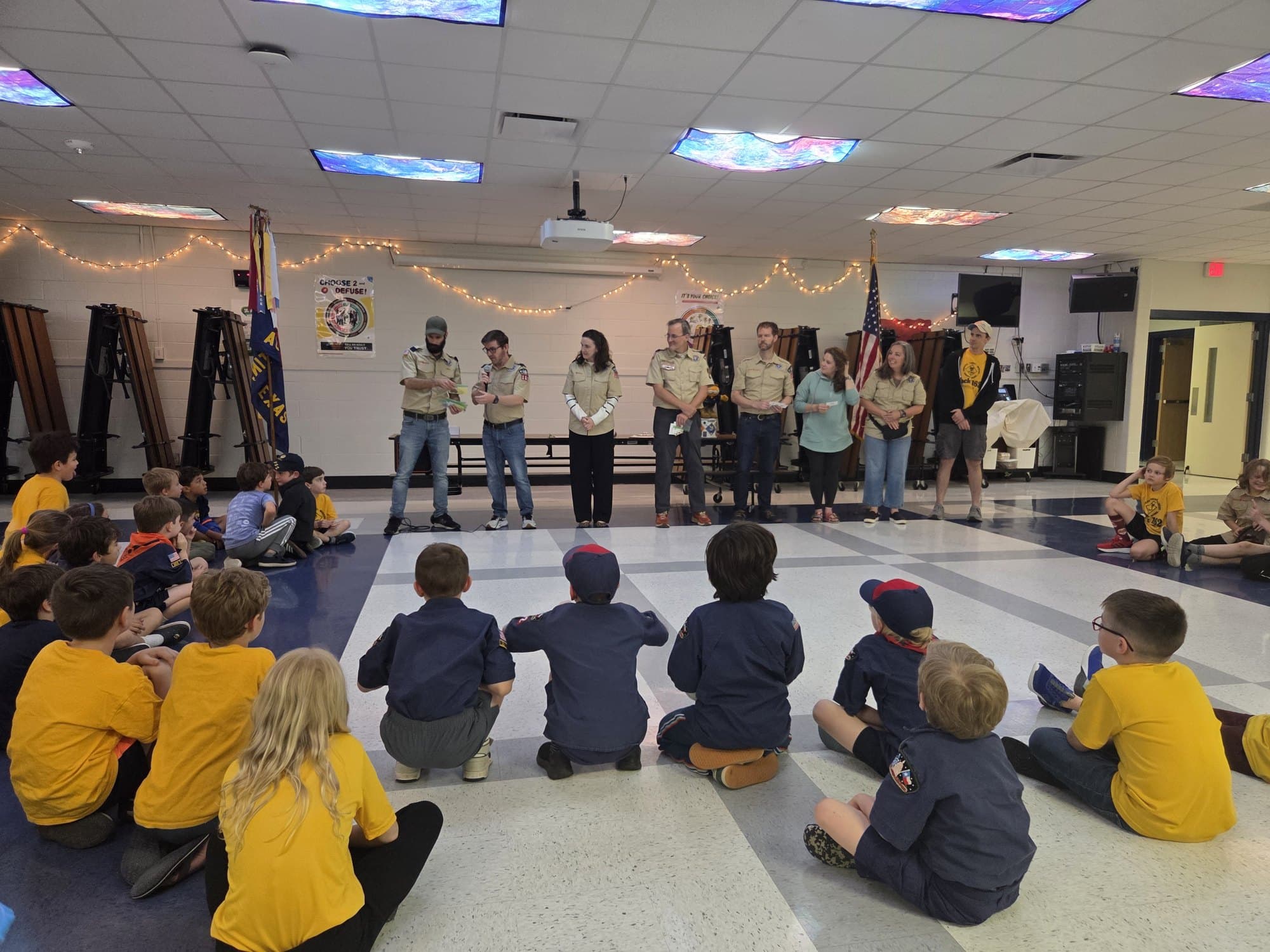 Pack 162 leaders standing together at the front of the school cafeteria, addressing scouts seated on the floor